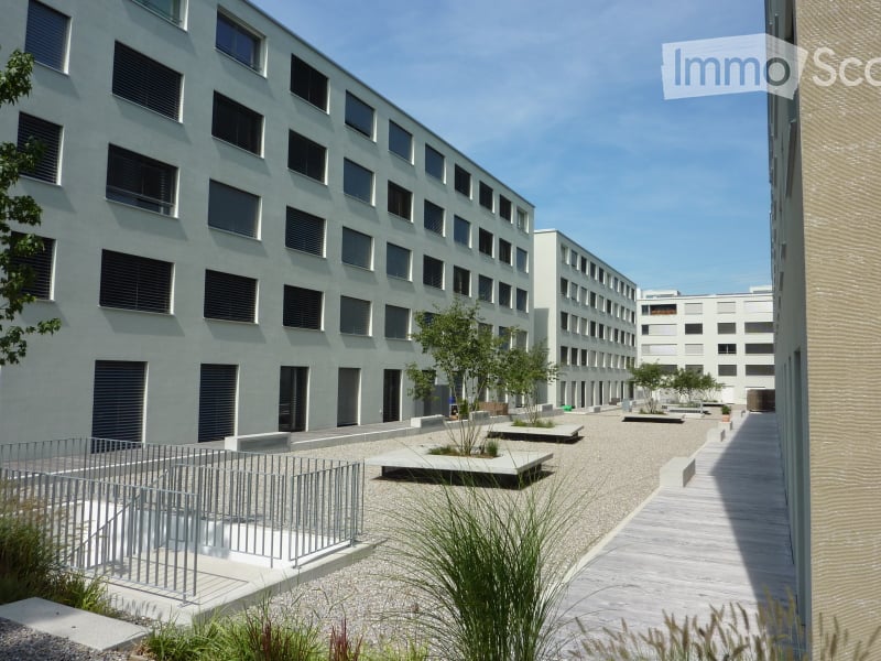 Modern multi-story building with a clean, minimalist design, featuring numerous windows and a raised ground floor with a paved courtyard and low seating areas.