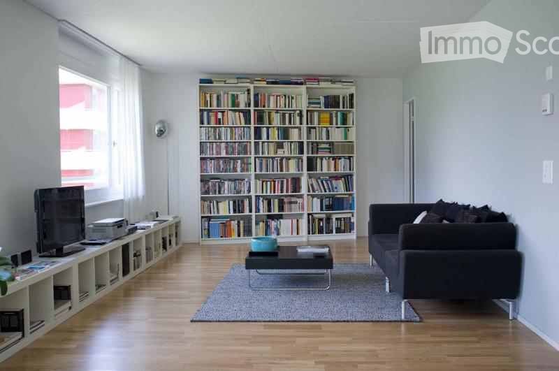 Modern living room with light grey walls, wooden flooring, large window with white curtains, black sofa, coffee table, bookshelf filled with books, and a television on a white stand.