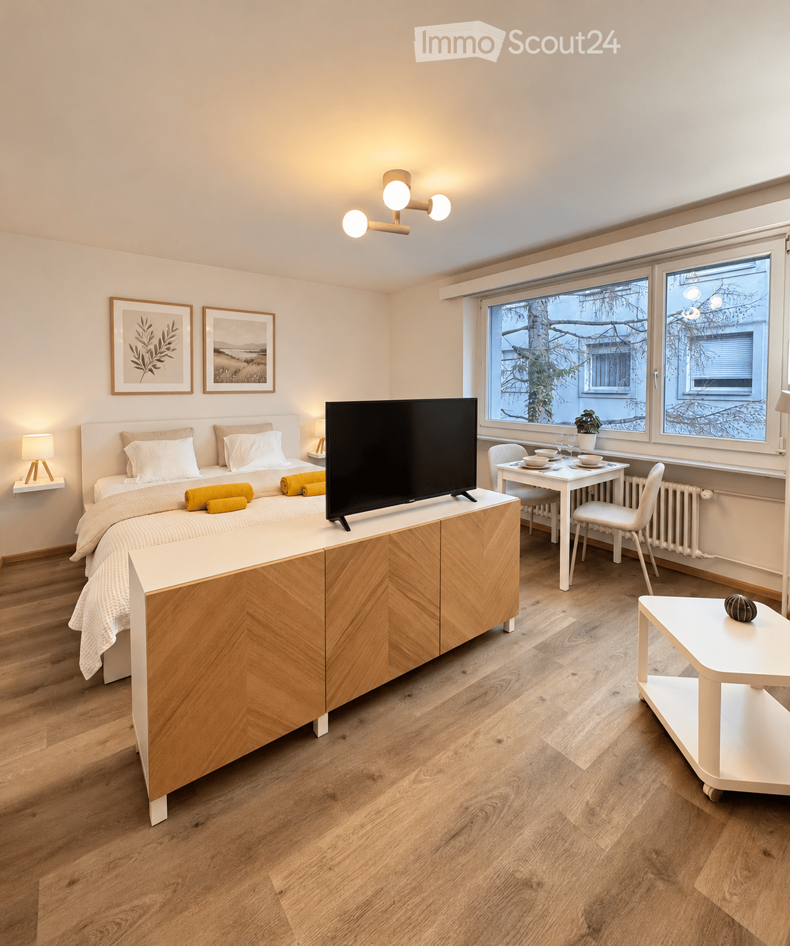 A modern, well-lit bedroom with a large bed, a television on a wooden cabinet, a small dining area with a table and chairs, and decorative elements like picture frames and lamps.