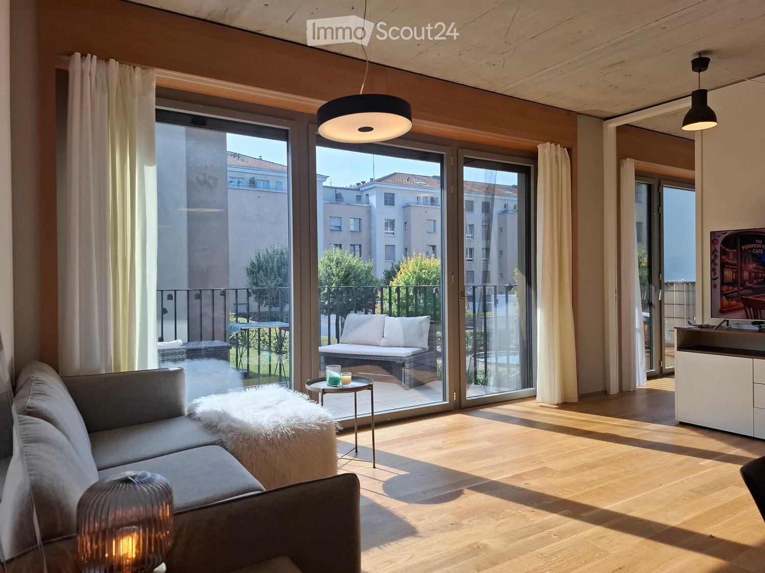 Modern living room with wooden flooring, large glass doors leading to a balcony, sofa, coffee table, ottoman, and a small table with drinks.