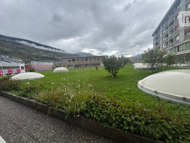A well-maintained garden area with green grass, small trees, shrubs, and several dome-shaped structures in the background, with a modern building and mountains visible in the distance.