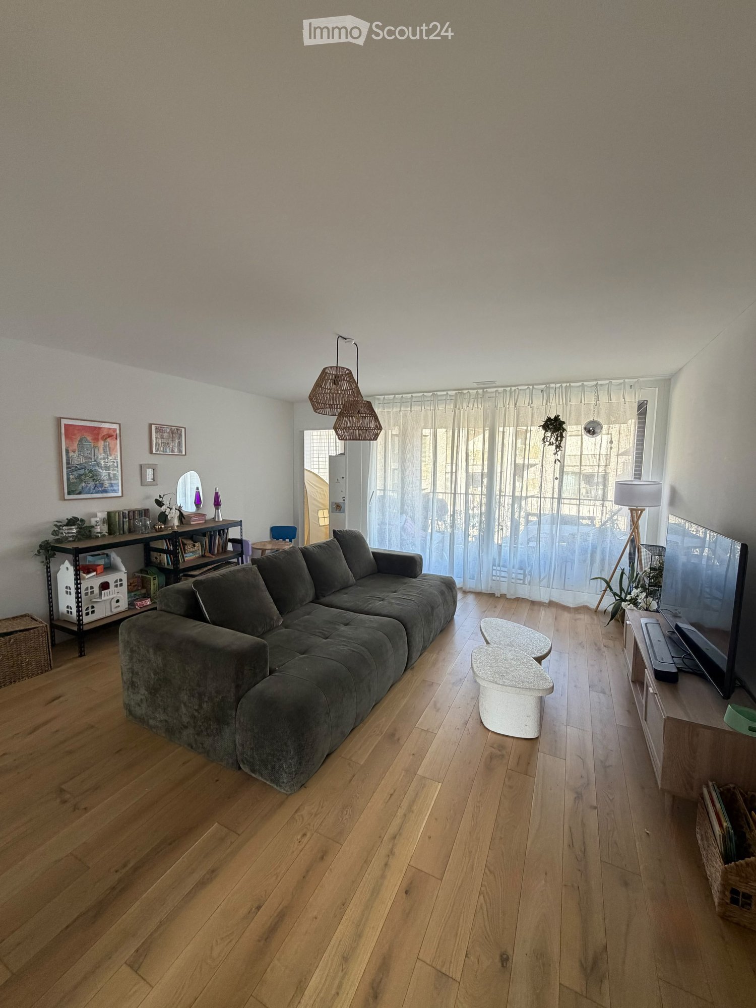 Modern living room with wooden flooring, large sectional sofa, coffee table, bookshelf, mirror, and television. Natural light from large windows with sheer curtains.