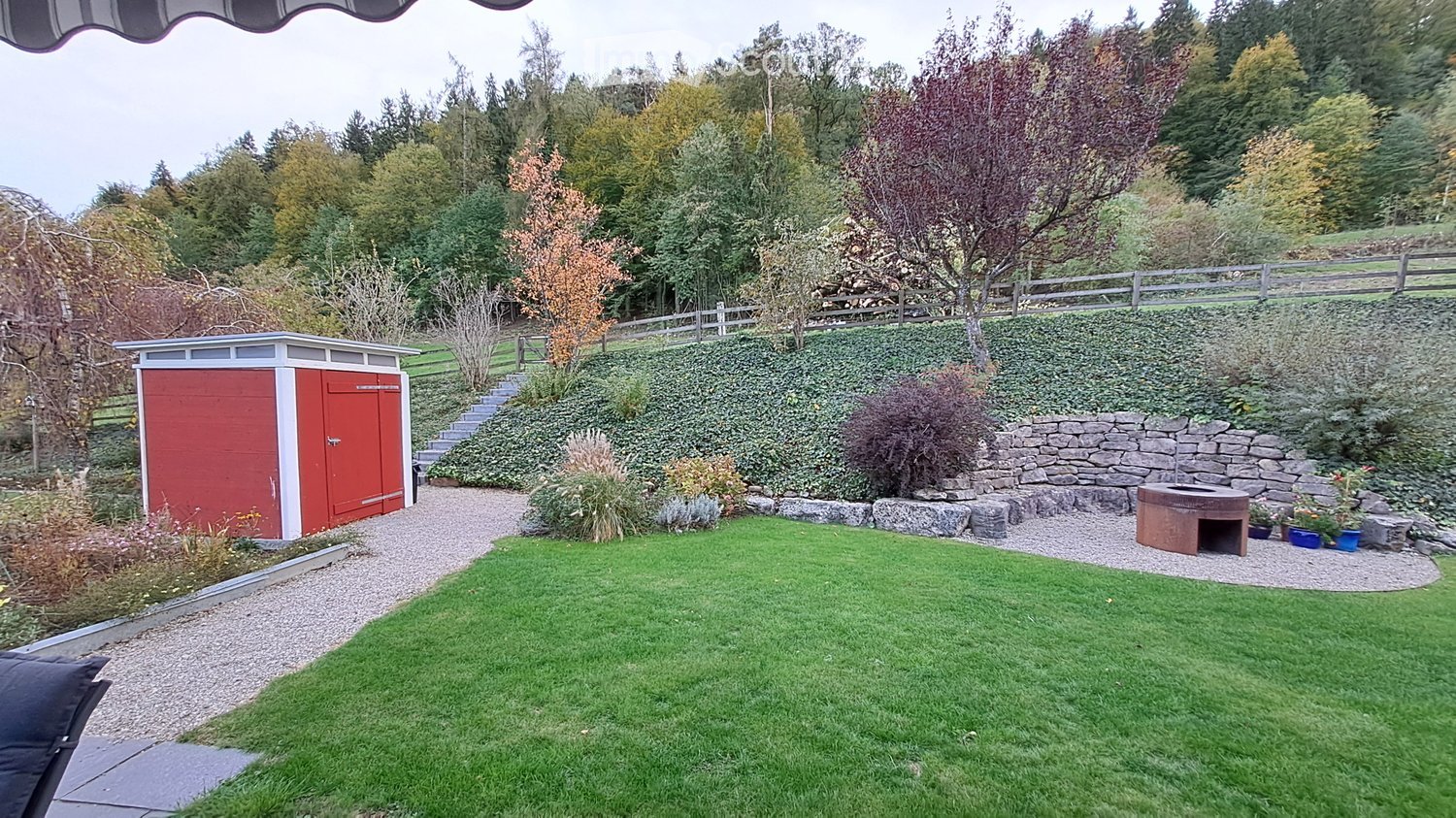 Well-maintained garden with a lawn, gravel pathways, a red shed, stone wall, and small seating area with a fire pit.
