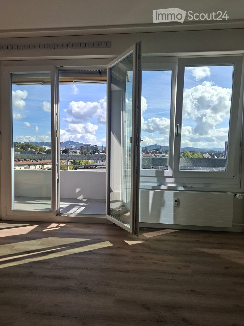 Empty room with wooden flooring, large windows, and open sliding doors leading to a balcony with a cityscape view.