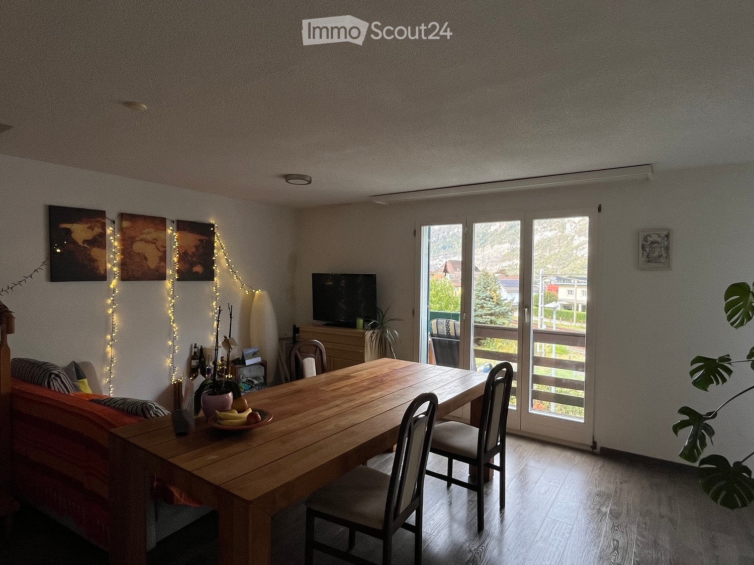 Bright living and dining area with wooden table, chairs, sofa, TV, and large windows leading to a balcony.