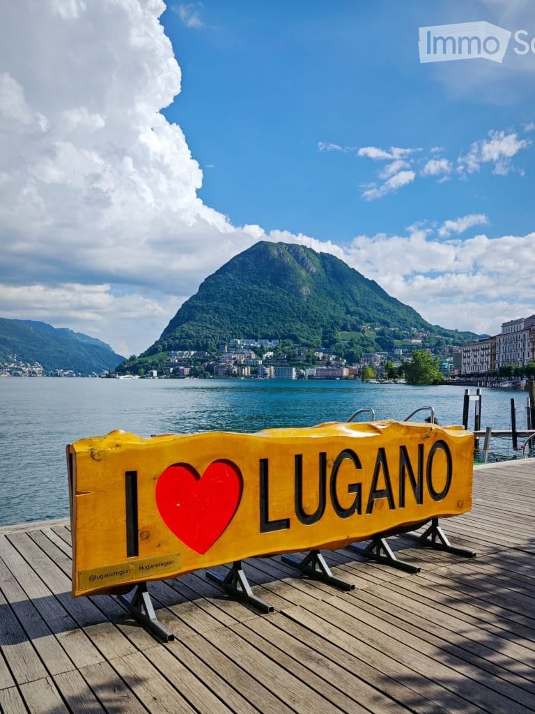 A scenic lakeside view with a wooden dock, a yellow sign reading "I LOVE LUGANO" with a red heart, and a distant mountain backdrop.