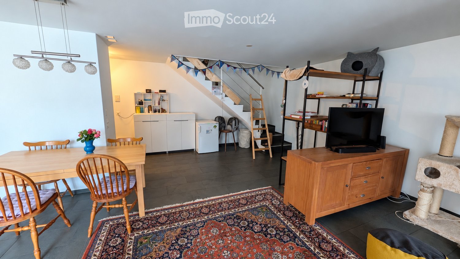 A well-lit, multi-functional room with a dining area, a TV area, and a small kitchen. Features include a wooden dining table with chairs, a TV on a wooden cabinet, a cat tree, a small refrigerator, and a staircase leading to another level.