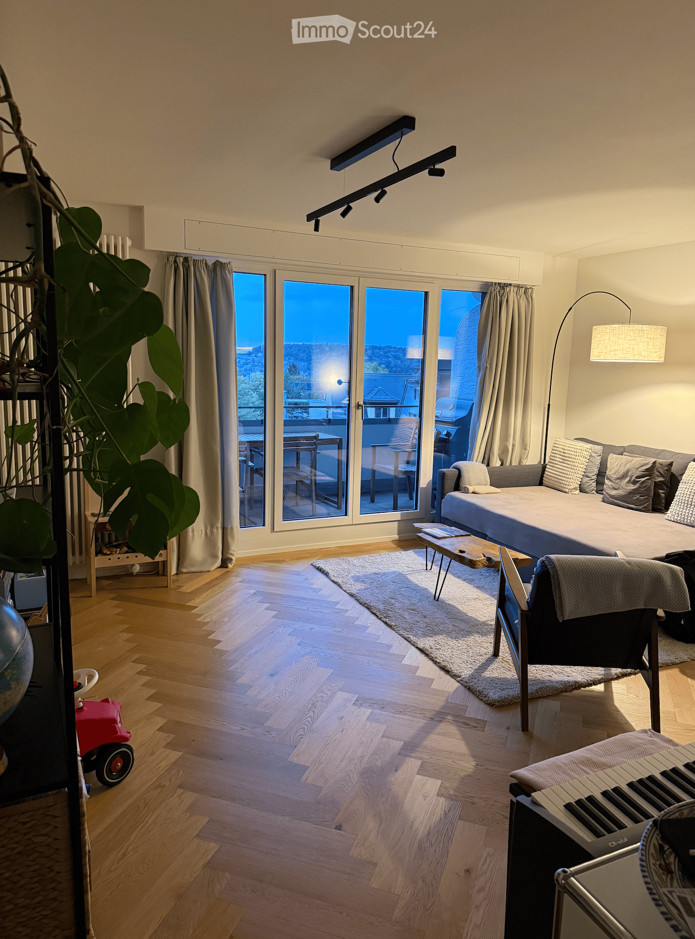 Modern living room with wooden flooring, large windows offering a view, a sofa, coffee table, chairs, and a small table on the balcony.
