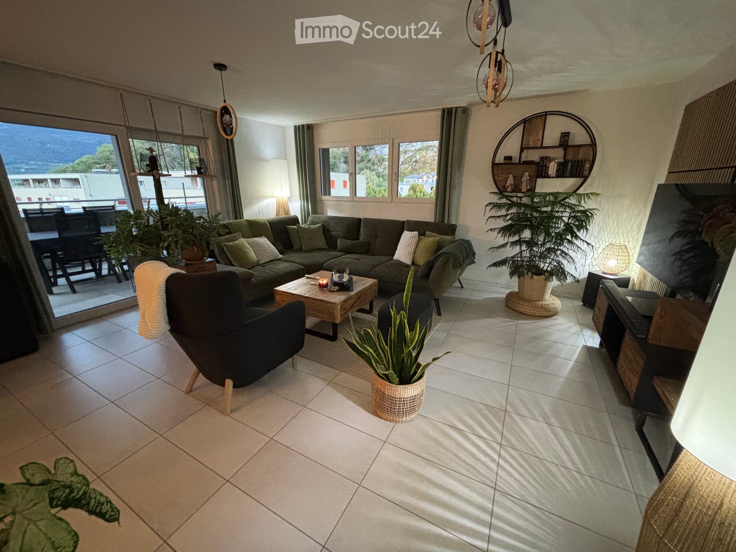 Modern living room with large windows offering a scenic view, featuring a sectional sofa, coffee table, armchair, and decorative plants.
