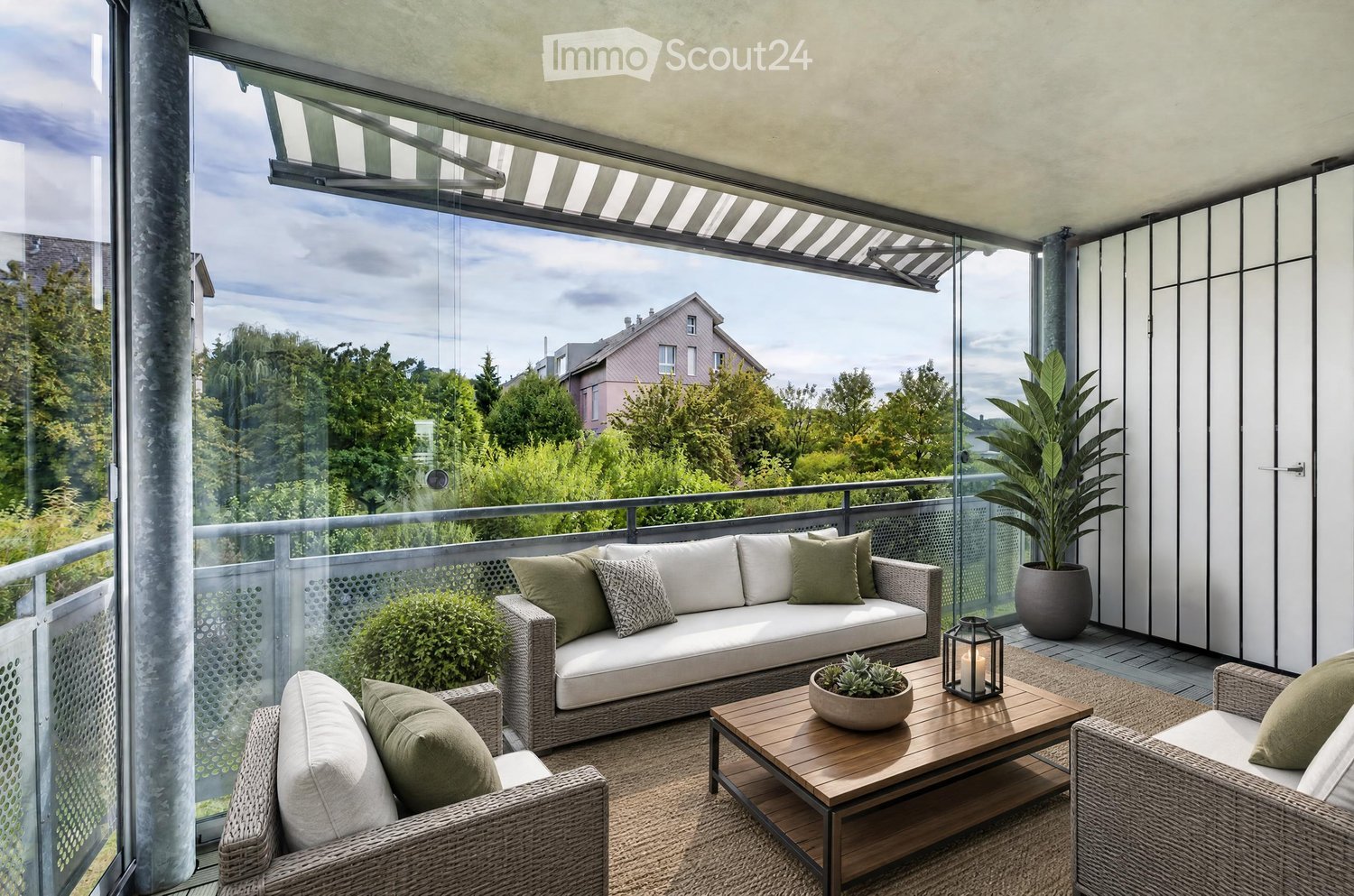 Modern balcony with large windows, offering a scenic view of trees and houses. Features a white wicker sofa, wooden coffee table, two wicker chairs, a potted plant, and a candle holder.