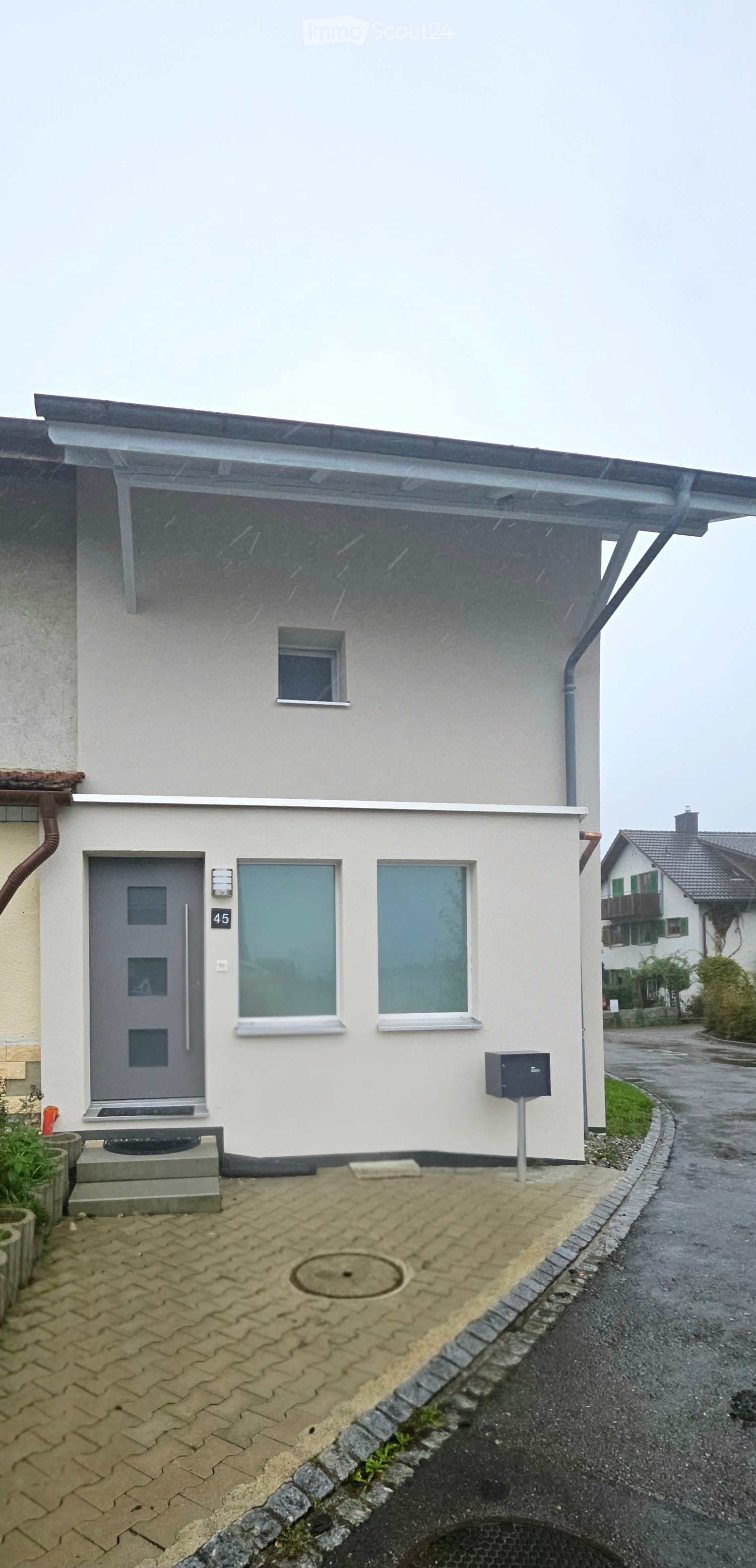 Single-story house with a gray front door, two windows, and a small porch area with a mailbox. The house has a modern design with a flat roof and a small garden area to the left.