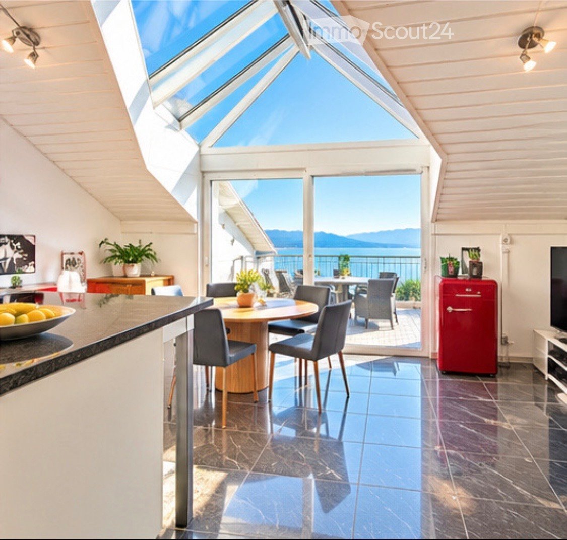 Bright, airy room with a skylight and large windows offering a scenic view. Features a round dining table with chairs, a red refrigerator, potted plants, and a bowl of fruit on the counter.