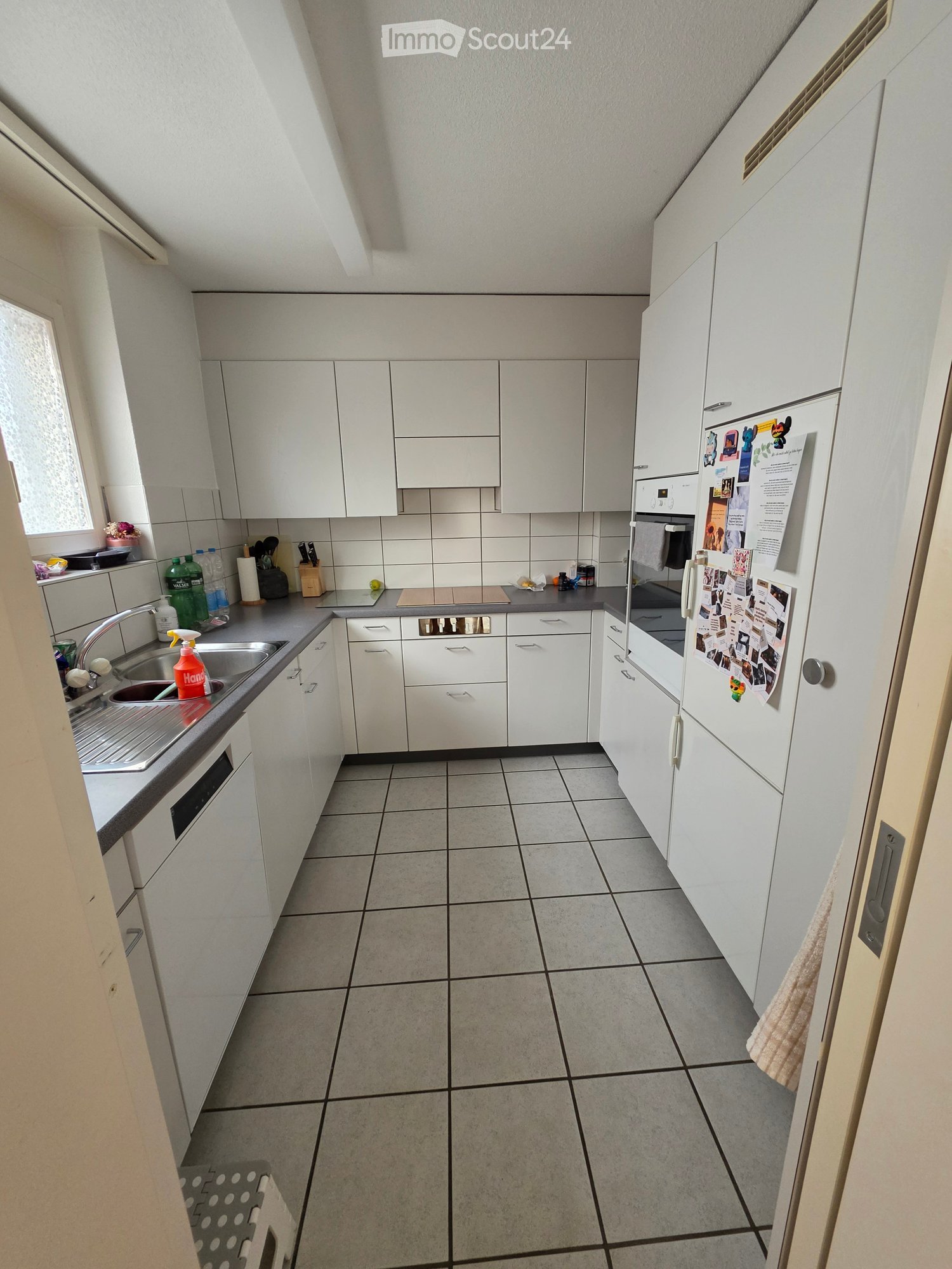 Modern kitchen with white cabinets, tiled floor, stainless steel sink, refrigerator, and countertop items including dish soap and cutting board.