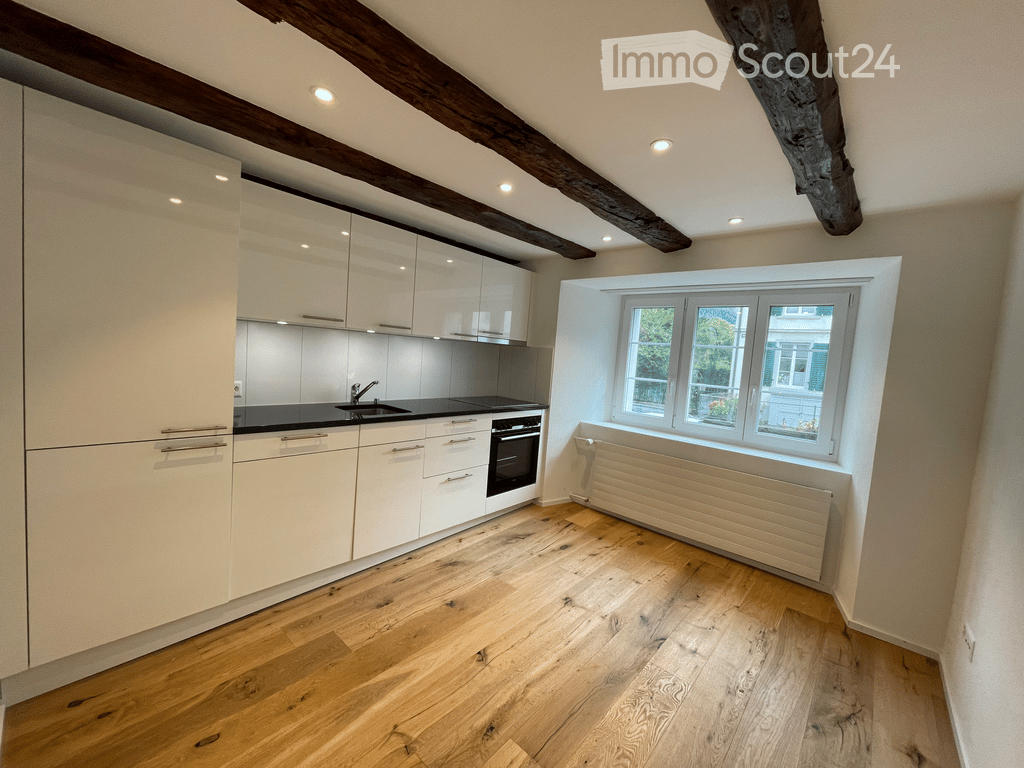 Modern kitchen with light-colored cabinets, a black countertop, and wooden flooring. Features include a sink, oven, and a window with a radiator below.