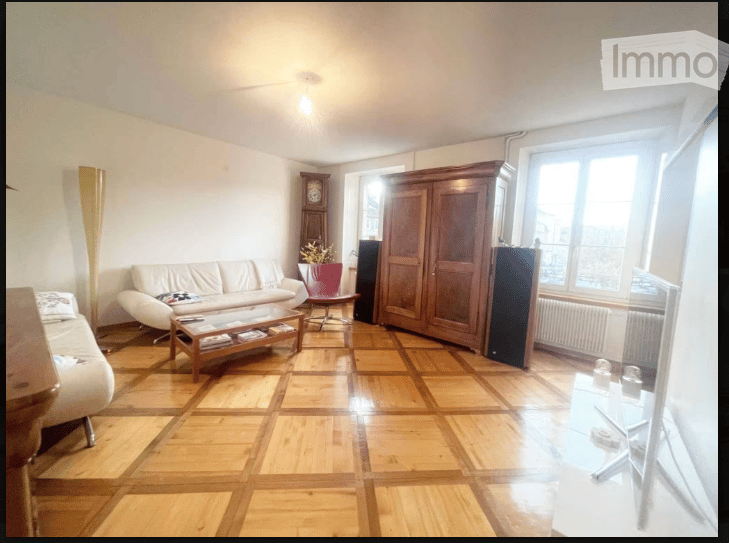 Living room with wooden parquet flooring, white walls, large window, sofa, coffee table, armchair, wardrobe, floor lamp, and television.