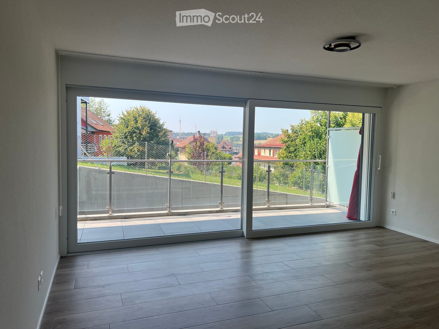 Empty room with wooden flooring, large sliding glass doors leading to a balcony with metal railings, and a view of greenery and buildings.