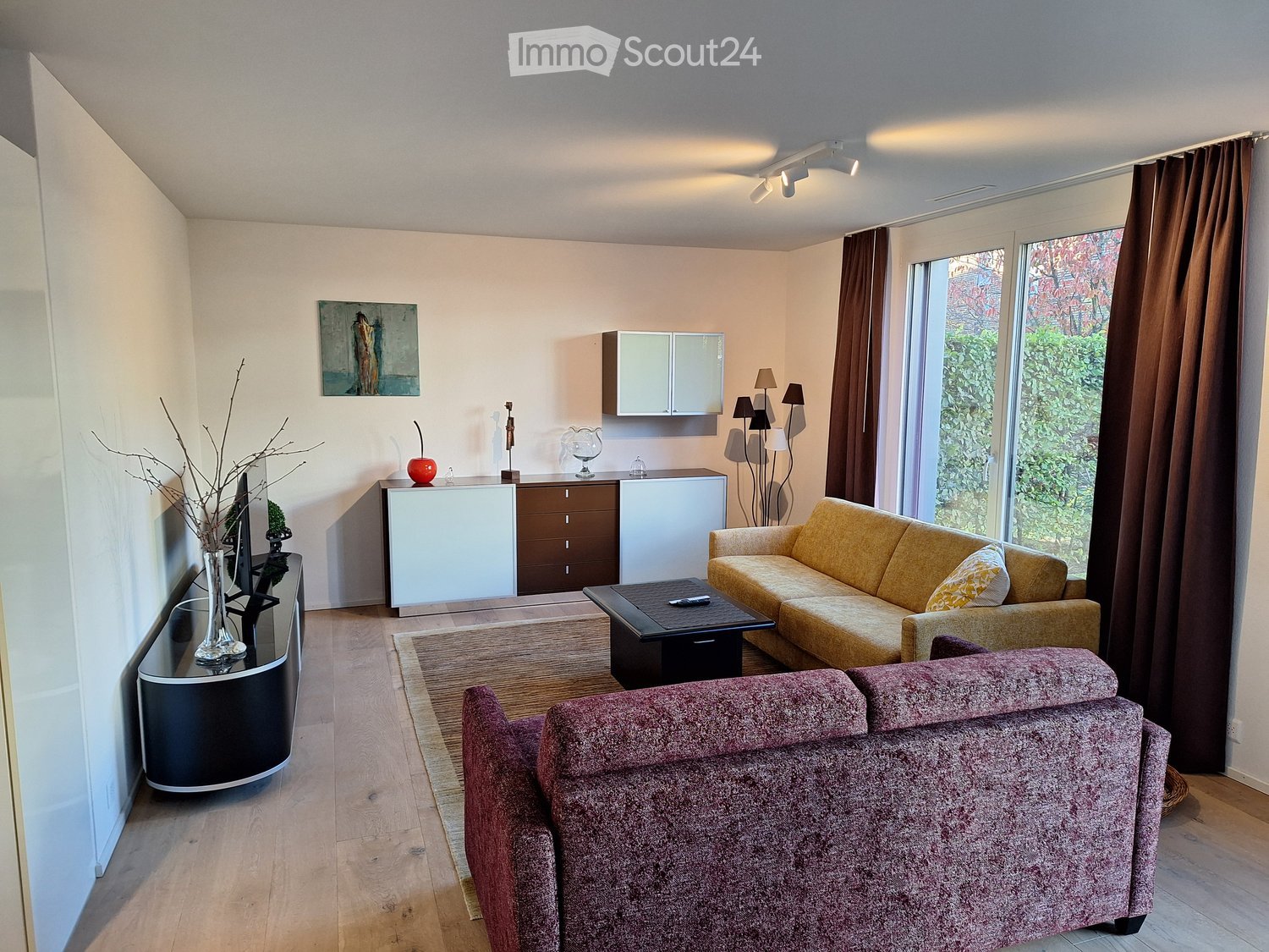 Modern living room with light wooden flooring, large windows with brown curtains, a yellow sofa, a red armchair, a black coffee table, a white cabinet, and decorative items including a vase with branches and a picture frame on the wall.