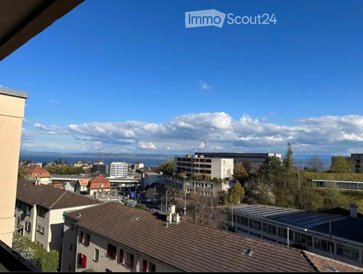 Panoramic cityscape view from a balcony, featuring rooftops, buildings, and distant water body under a clear blue sky.