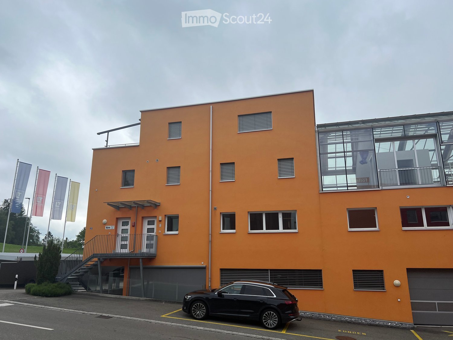Modern two-story building with orange facade, large windows, and a glass balcony area on the upper floor. Features include a staircase leading to the entrance, a parked car in front, and several flags on poles to the left.