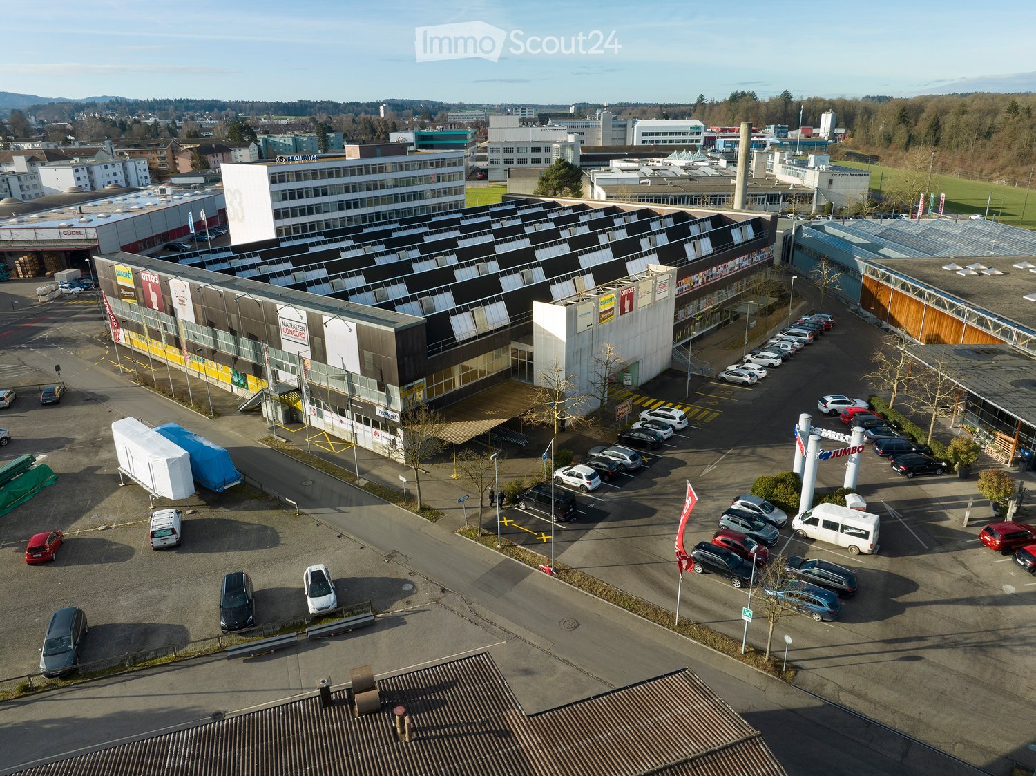 Aerial view of a commercial building complex with multiple storefronts, parking lot, and surrounding urban area.