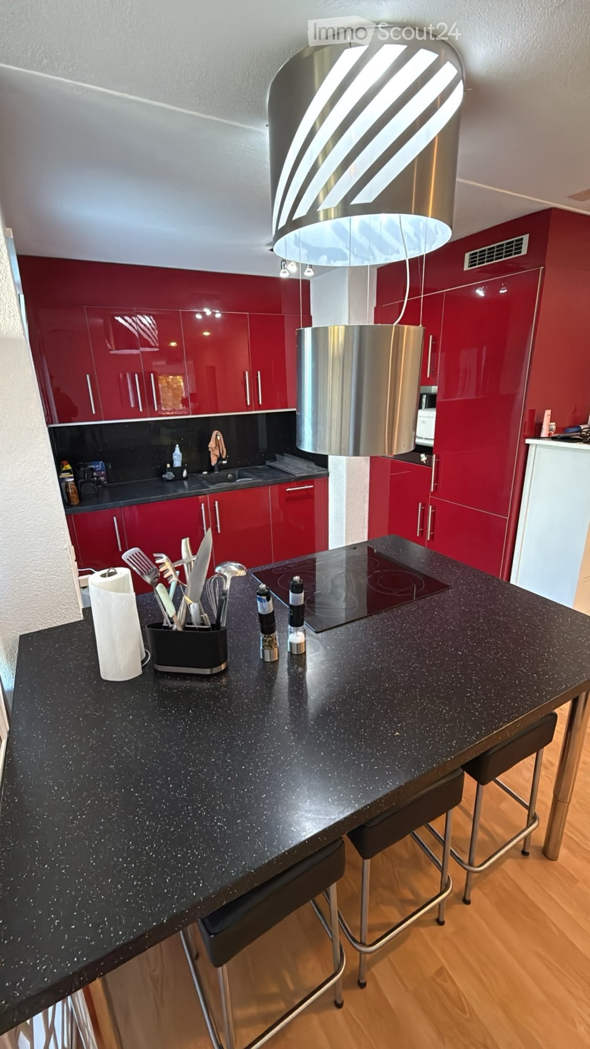 Modern kitchen with red cabinets, black countertop, induction cooktop, and a hanging pendant light. Kitchen utensils and a paper towel are on the counter.