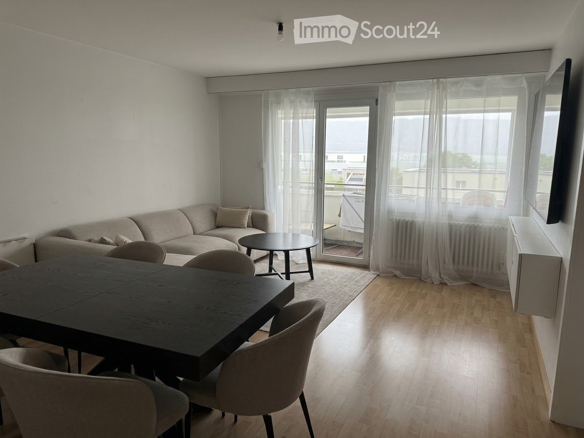 Modern living and dining area with a light-colored sofa, coffee table, dining table, and chairs. Large windows and a sliding glass door lead to a balcony.