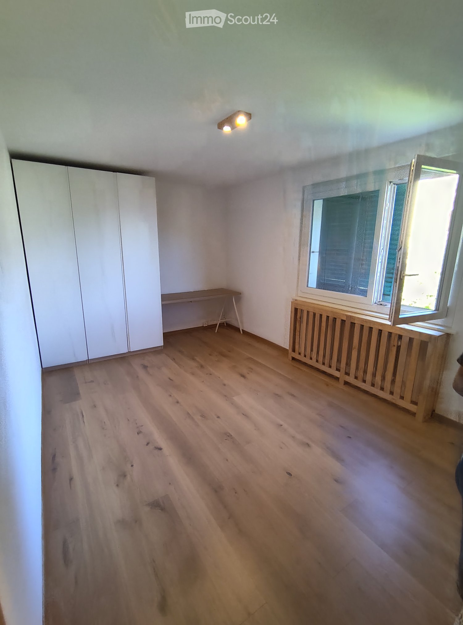 Empty room with wooden flooring, white walls, a window with blinds, a small wooden table, and a built-in wardrobe.