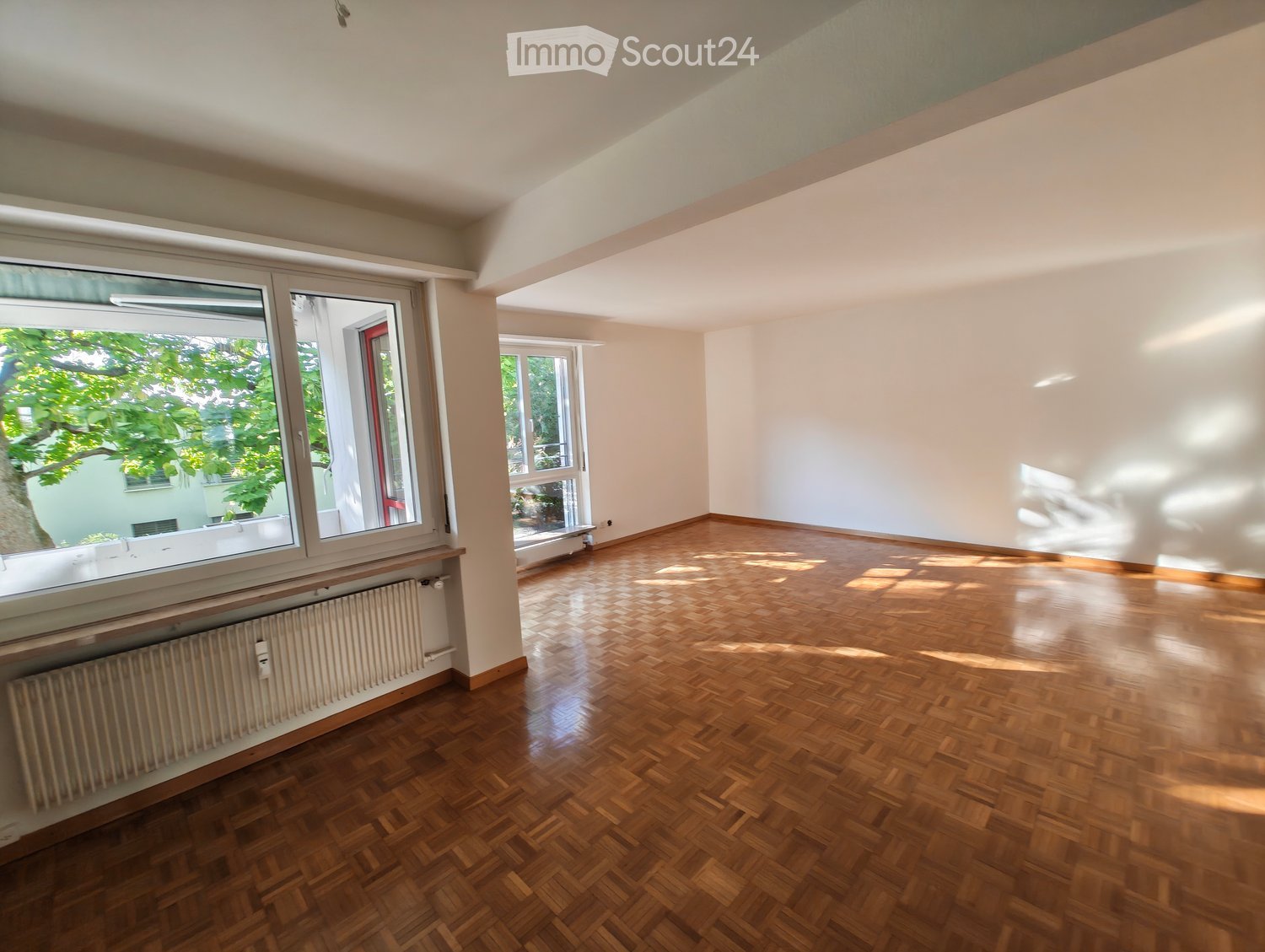 Empty room with wooden parquet flooring, white walls, large windows, and a radiator under one window.