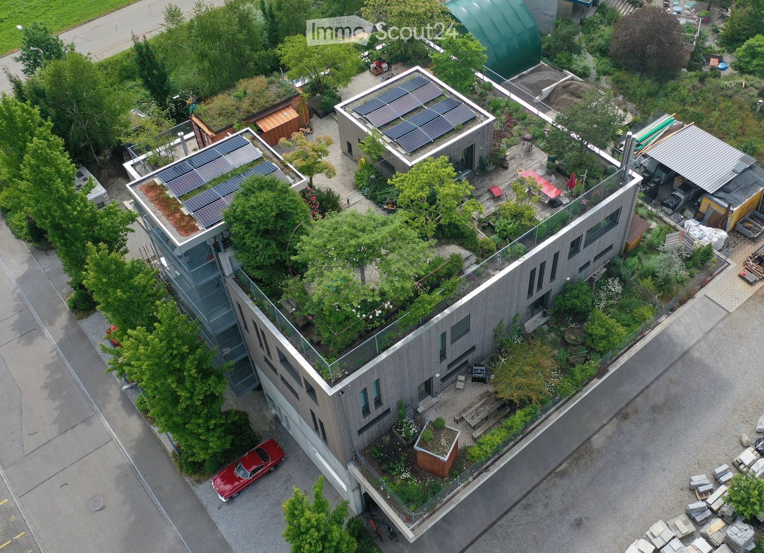 A modern, multi-story building with a green roof, solar panels, and surrounding greenery. Features include a staircase leading to the roof, outdoor seating, and a small garden area.