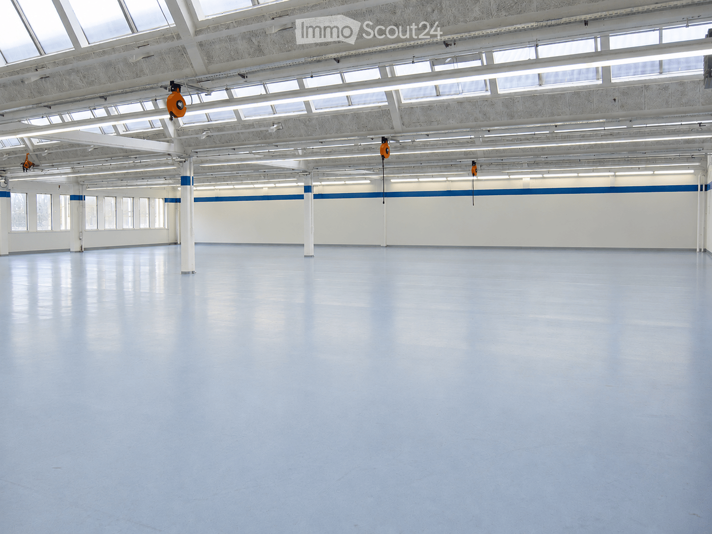 Large, empty industrial-style room with white walls, polished blue floor, and exposed white structural beams and lighting. No furniture or equipment present.
