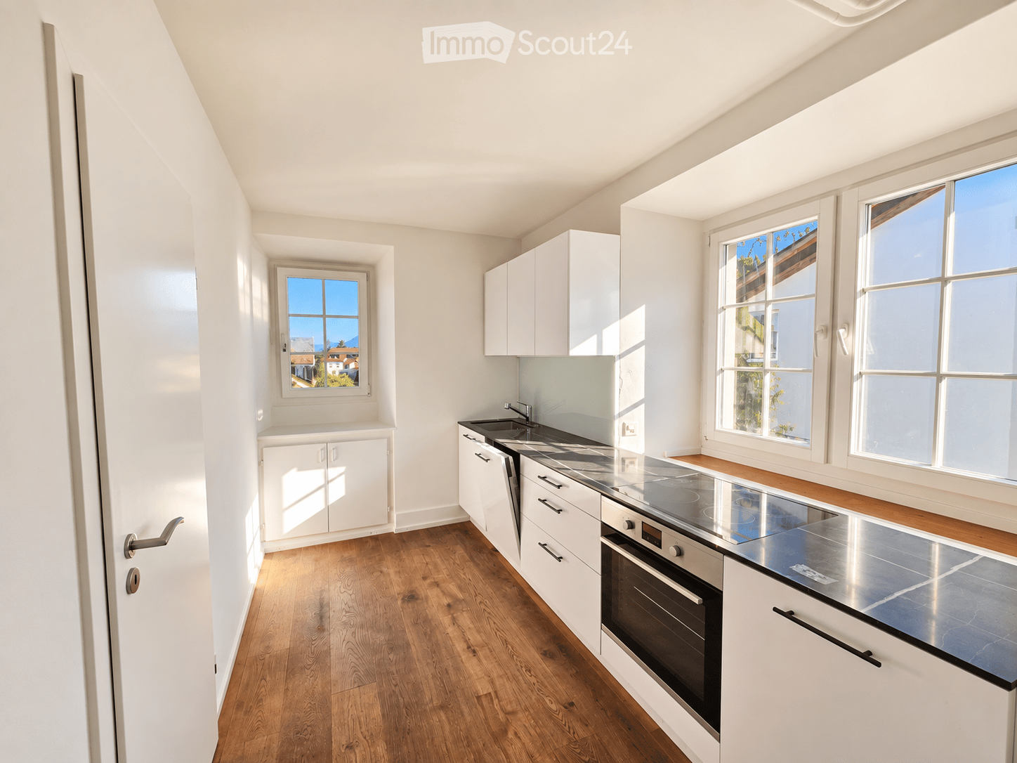 Modern kitchen with white cabinets, stainless steel appliances, wooden flooring, and large windows offering a view.
