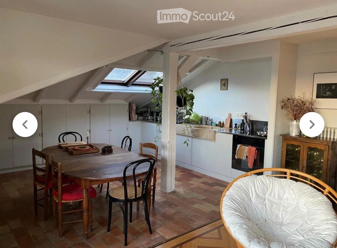 A cozy attic space with a dining area and a small kitchen. The dining area features a wooden table with chairs, and the kitchen has white cabinets, a sink, and an oven. The room has exposed wooden beams and skylights, creating a bright and airy atmosphere