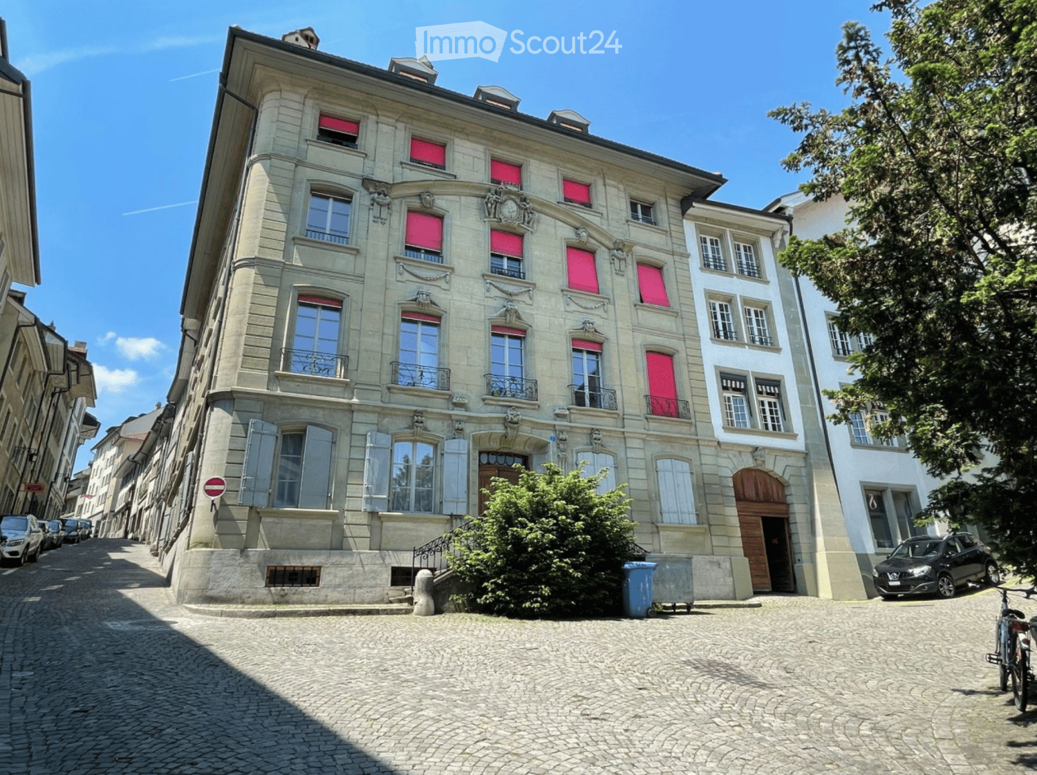 A multi-story building with classical architecture, large windows, and red shutters, situated on a cobblestone street.