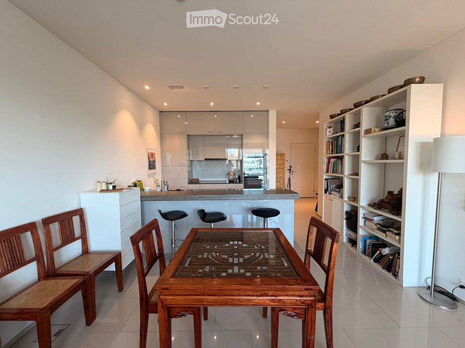 Bright, modern living and dining area with a wooden dining table, four wooden chairs, four black stools at a kitchen island, white cabinets, and built-in shelving with books and decorative items.
