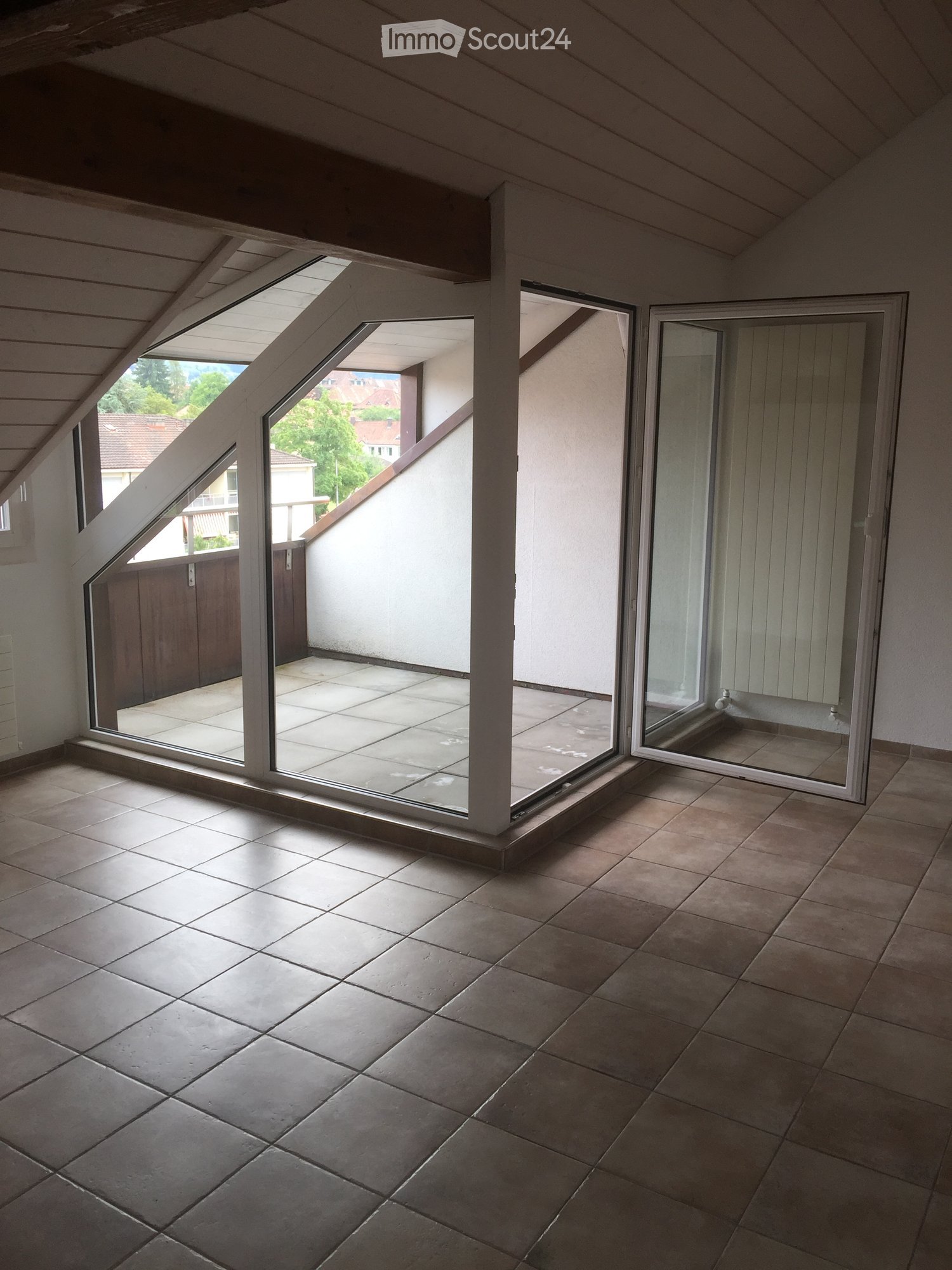 Empty room with tiled floor and large windows opening to a balcony area. The room has a slanted ceiling typical of an attic space.