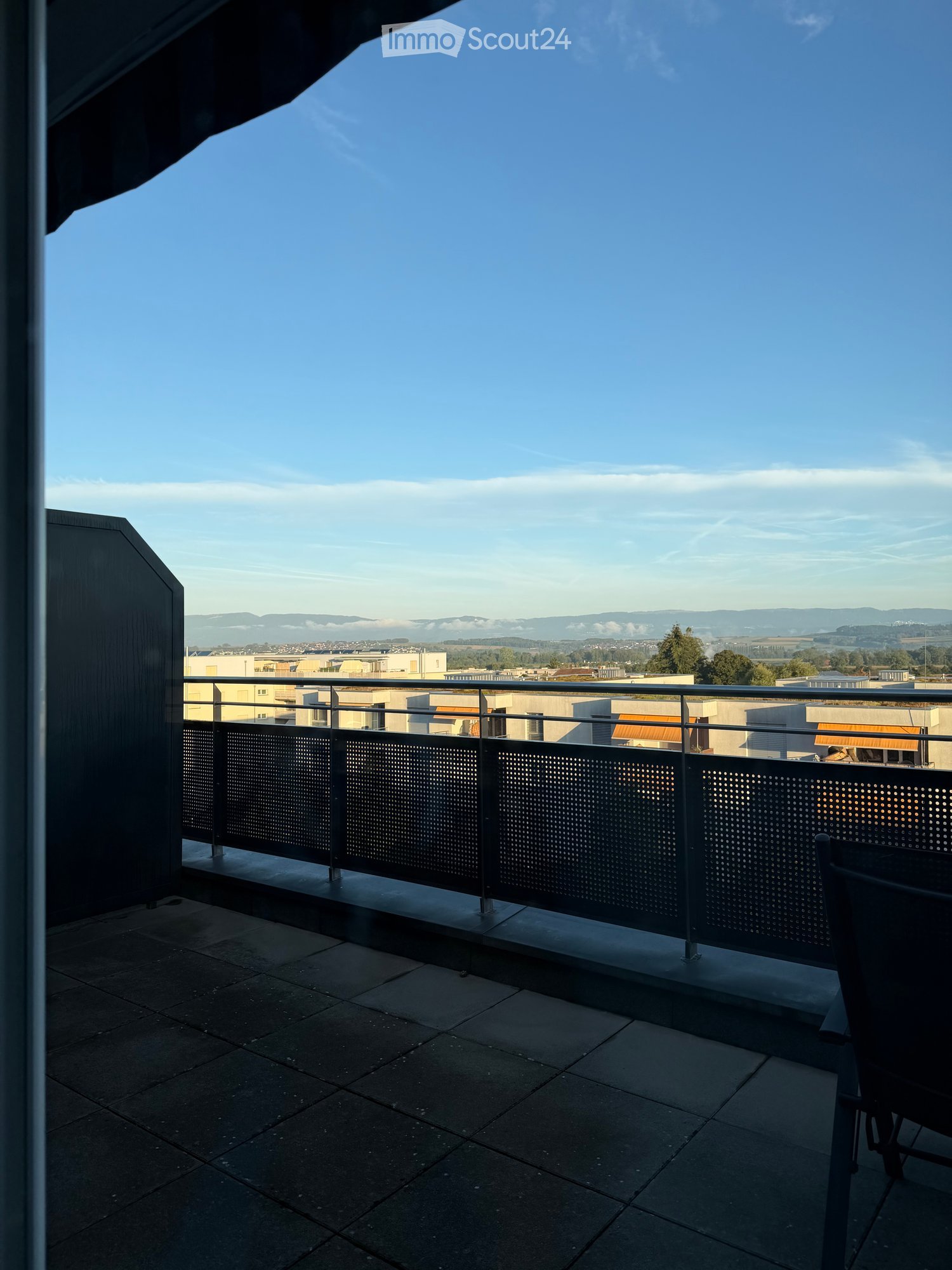 Balcony with a modern metal railing, tiled floor, and a scenic view of a cityscape and distant hills under a clear blue sky.