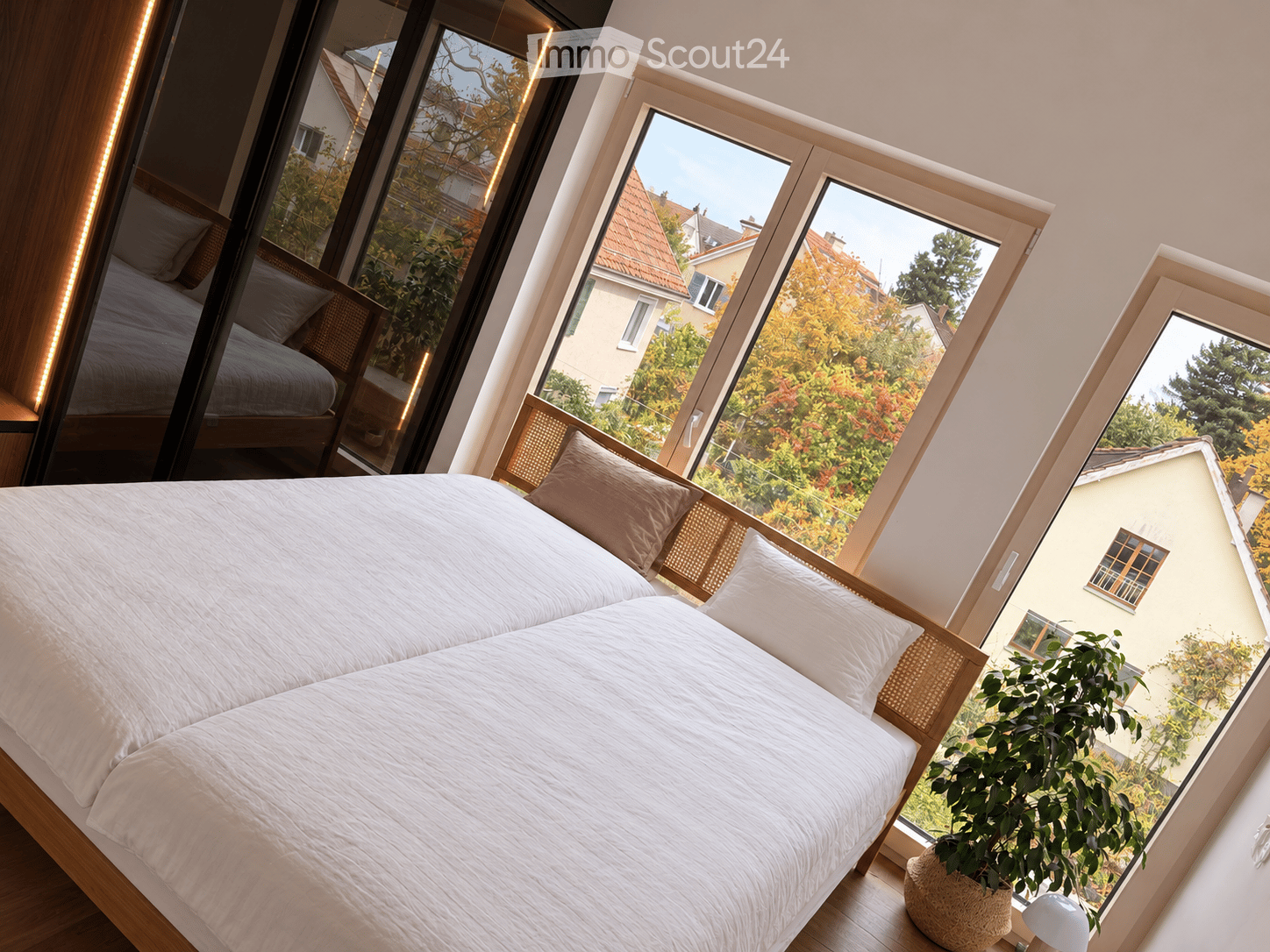 Modern bedroom with large windows offering a view of the garden and neighboring houses. Features a double bed with white bedding, a nightstand with a lamp, and a potted plant near the window.
