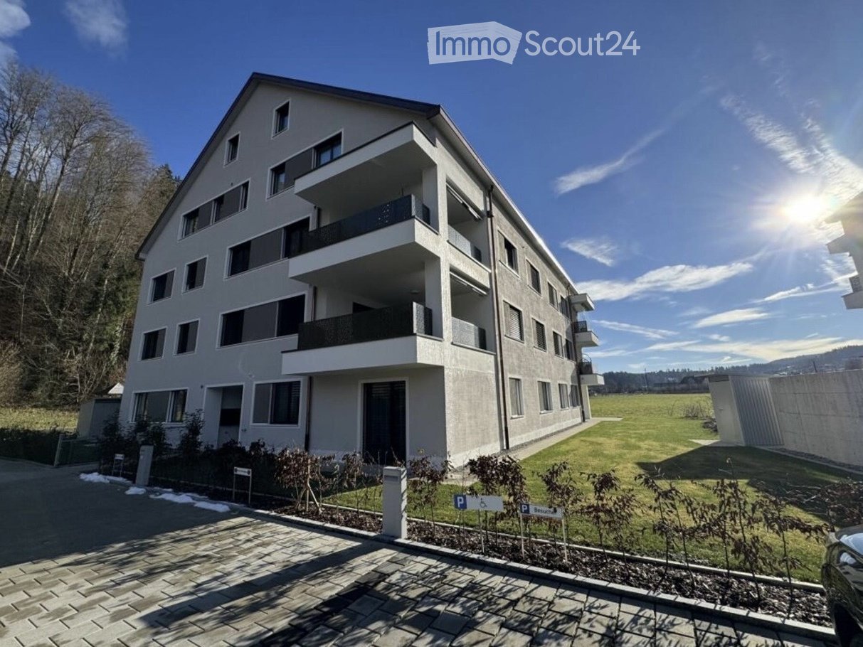 Modern multi-story residential building with balconies, parking area, and landscaped surroundings.