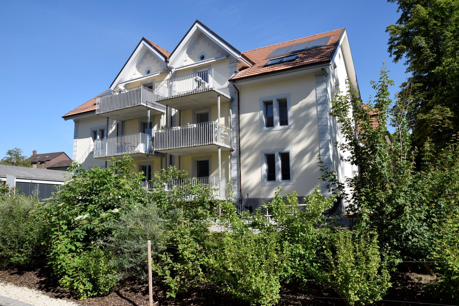 two-story building with balconies, solar panels on the roof, surrounded by plants