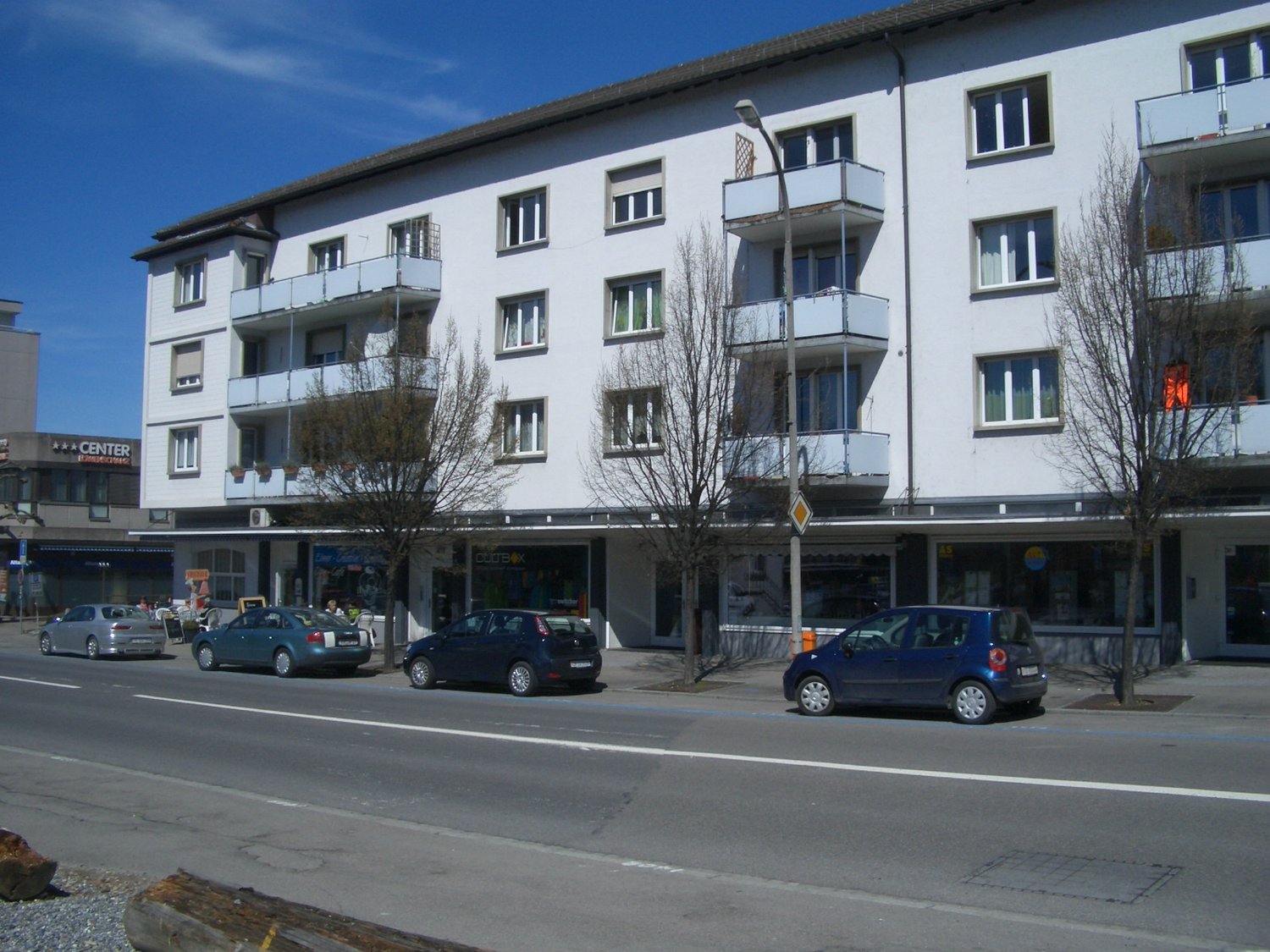 white building, multiple balconies, several floors, air conditioning unit, large windows, sidewalk, parking lot