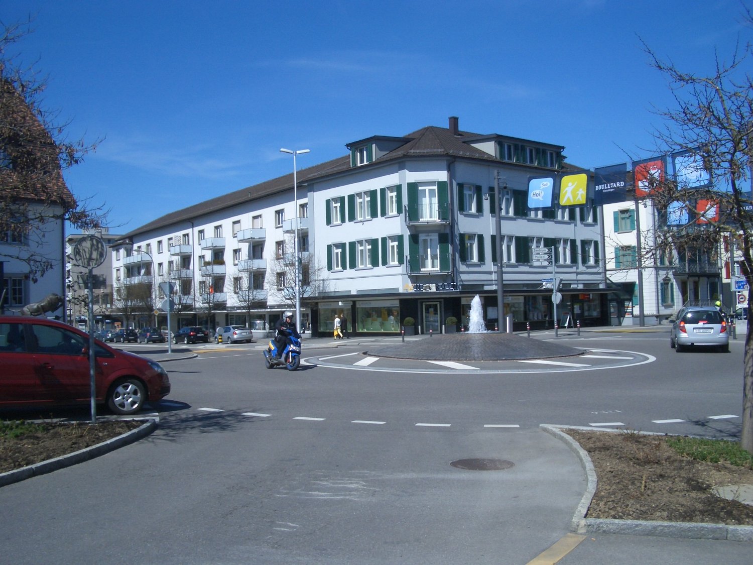 A wide intersection with an apartment building, fountain, streetlights, cars, a person on a scooter, and a tree.