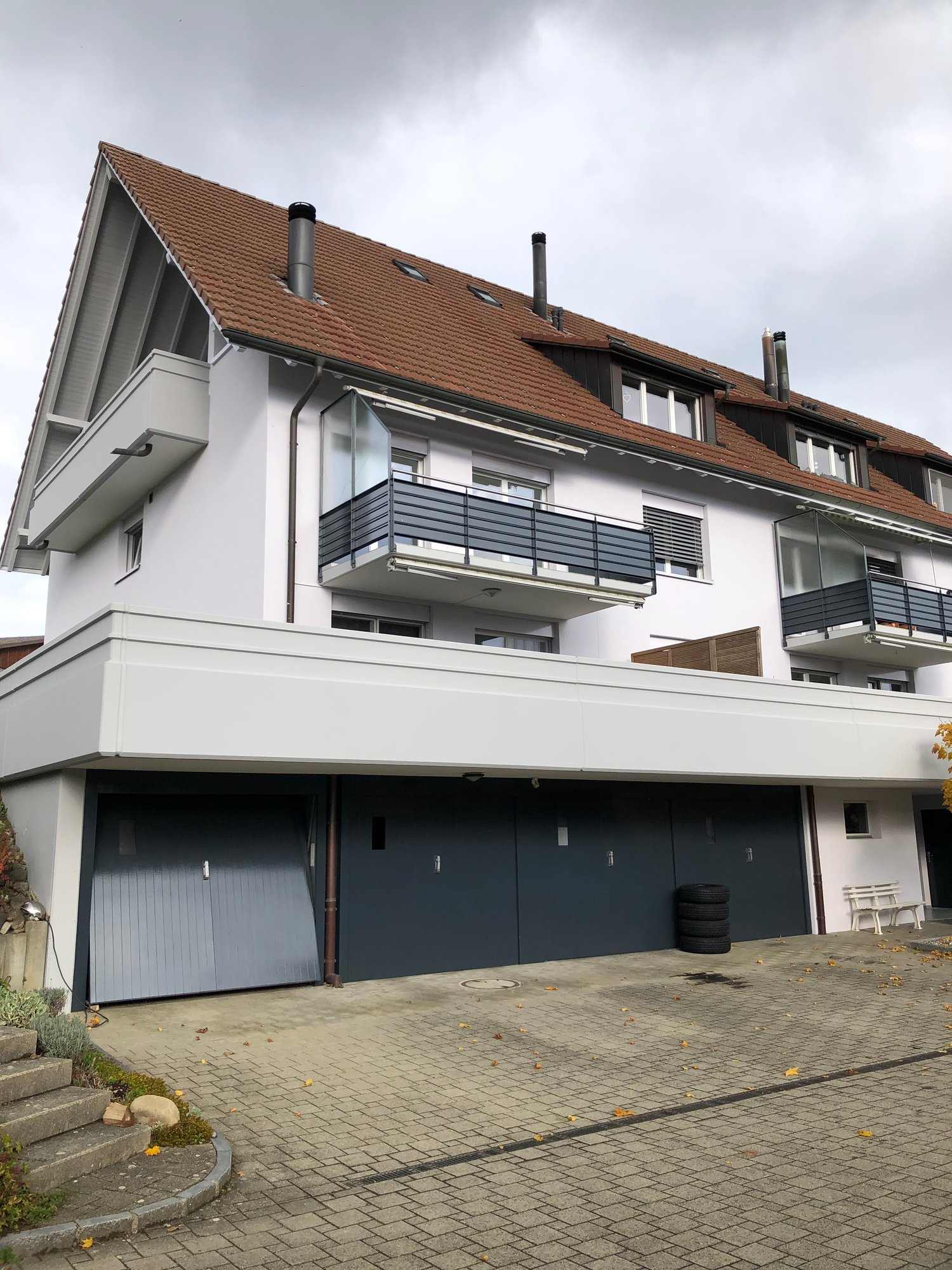Two-story white house with brown roof, balconies, garages in front, driveway with brick pavement, chimneys, and steps.
