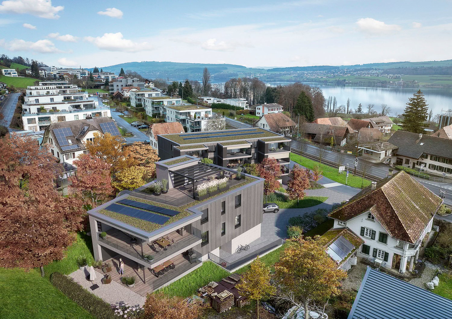 The image shows a scenic residential area overlooking a lake. The buildings have modern, multi-level designs with balconies and terraces. The area is surrounded by lush greenery and trees, creating a picturesque landscape. Solar panels can be seen on some