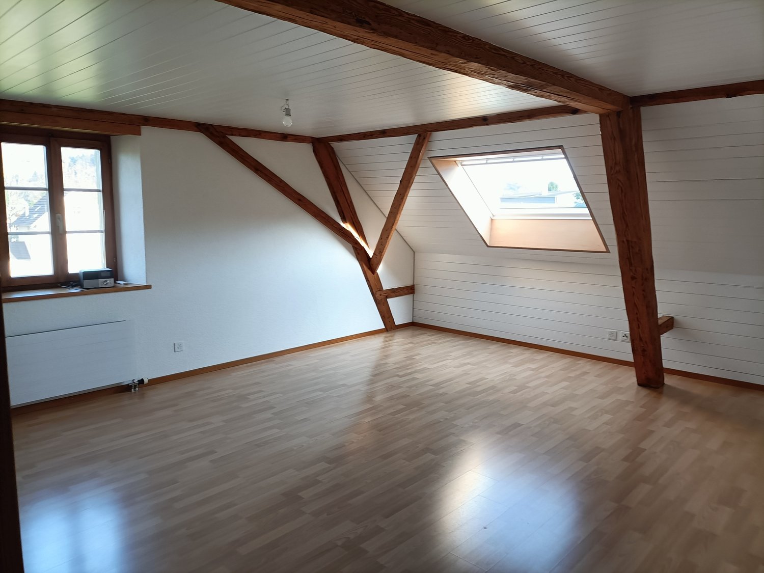 Empty room with wooden beams, wooden floor, white walls, window with wooden frame, wooden ceiling with attic window.