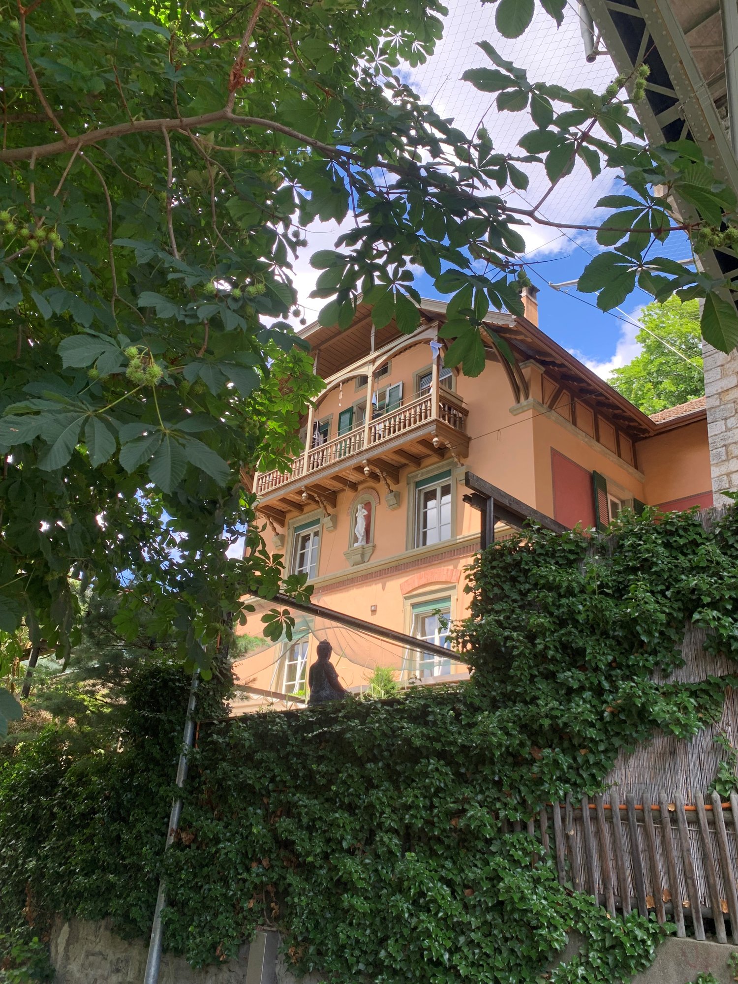 A multi-story building with a wooden balcony, surrounded by lush greenery and trees. The building has a traditional architectural style with decorative elements.