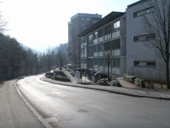 A white building with multiple windows, street light, greenery, and a pathway leading to the entrance.