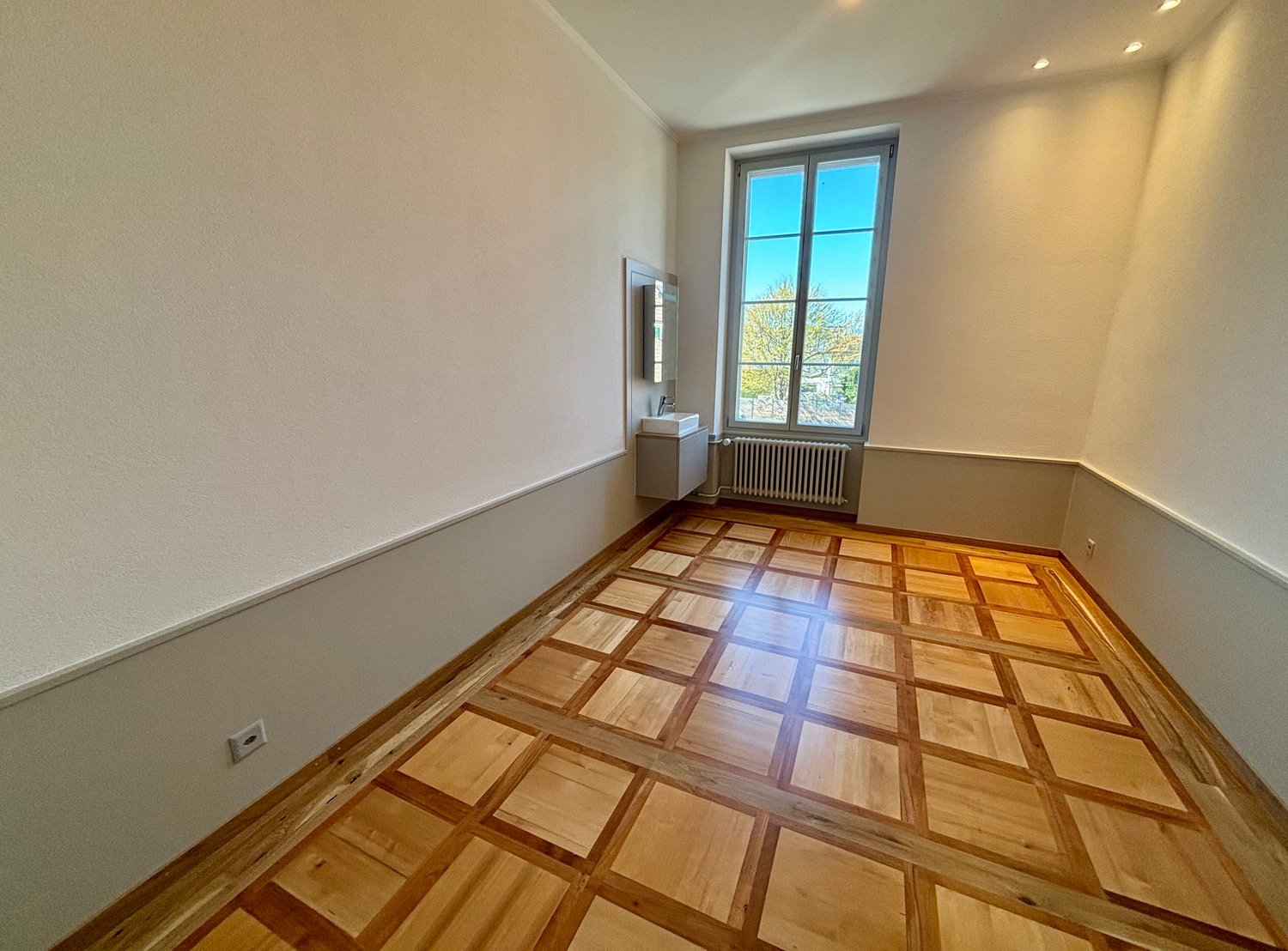 Empty room with wooden parquet flooring, white walls, a window, a small sink, and a radiator.