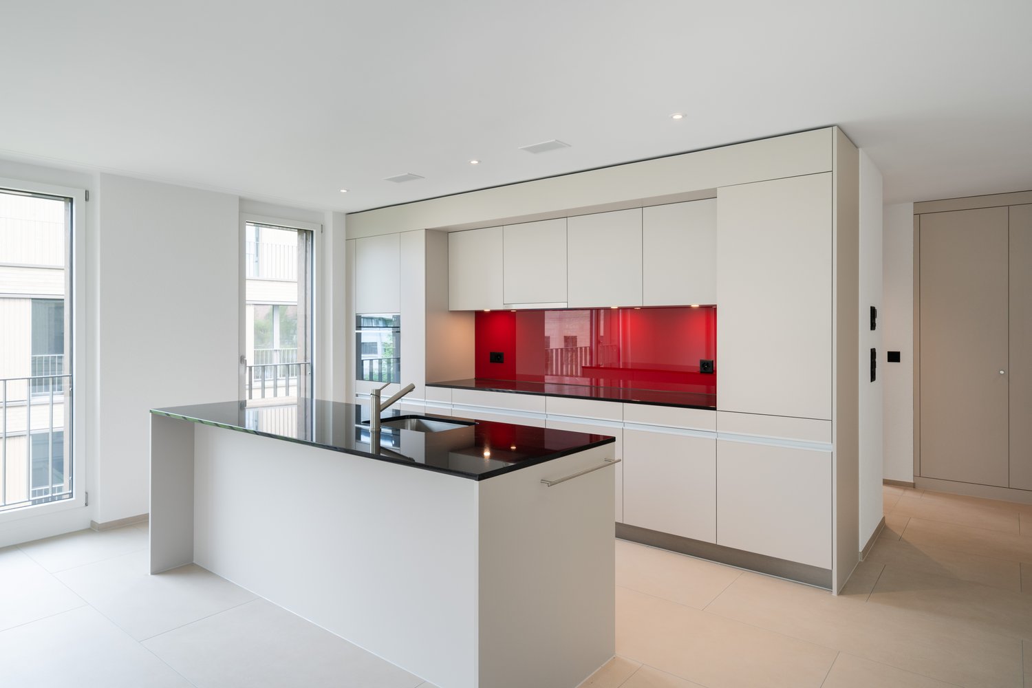Modern, open-concept kitchen with white cabinets, red glass backsplash, black countertop, and built-in appliances including a dishwasher. Large windows provide natural light and access to a balcony or terrace.