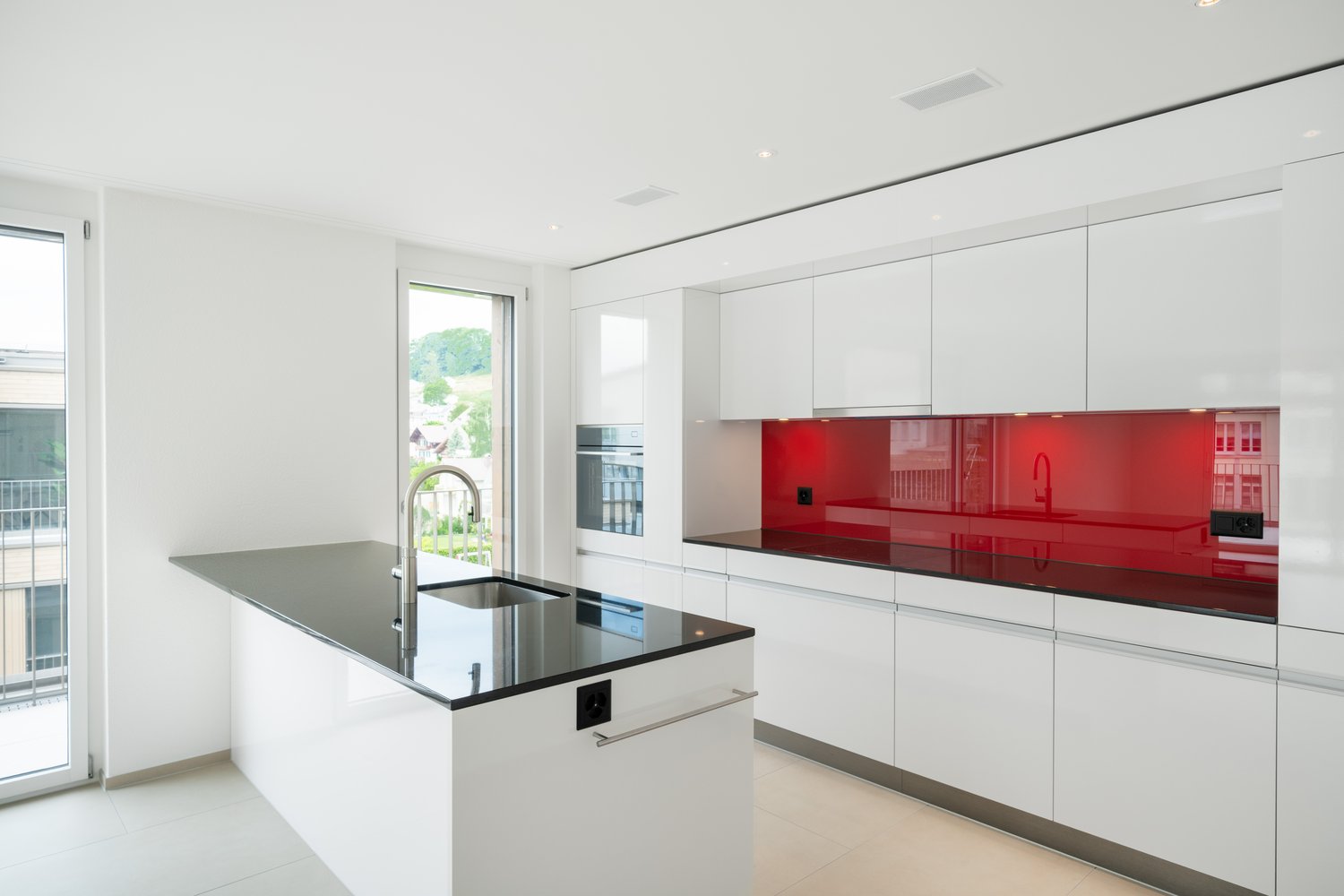 Modern, open-concept kitchen with white cabinets, red glass backsplash, and a large island with a black granite countertop. The kitchen features built-in appliances, including a dishwasher.