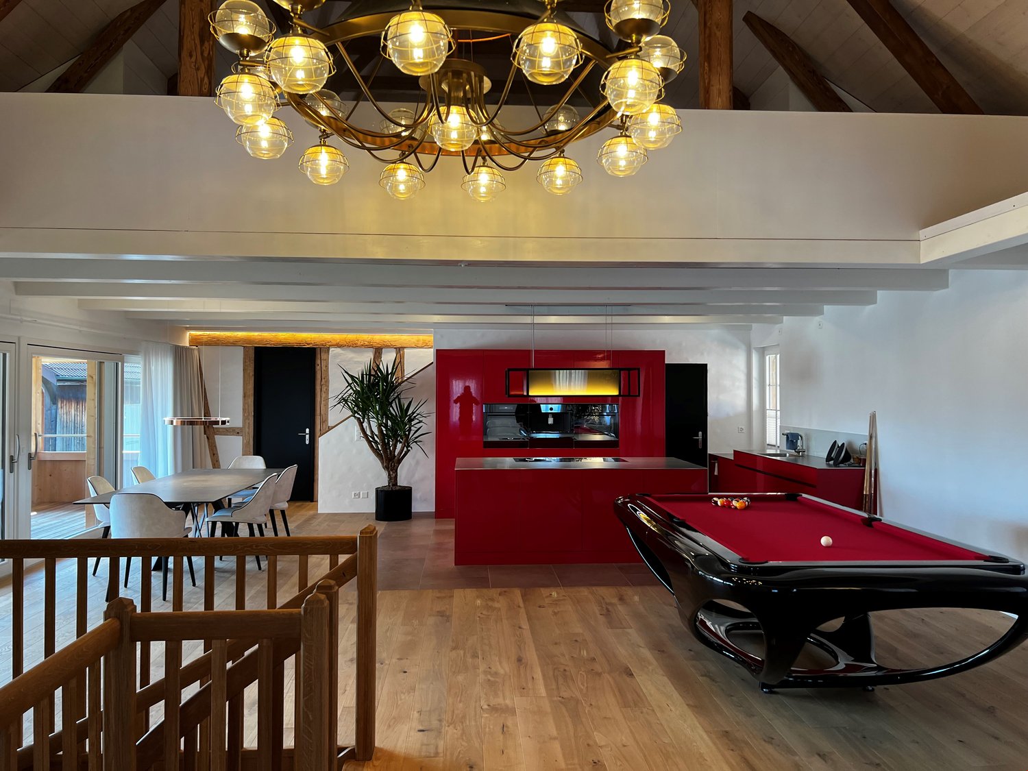 The image shows a large, open-concept living space with a dining area, kitchen, and entertainment area. The space features a high, vaulted ceiling with a large, ornate chandelier. The walls are a mix of white and red, with wooden beams and accents. The ki